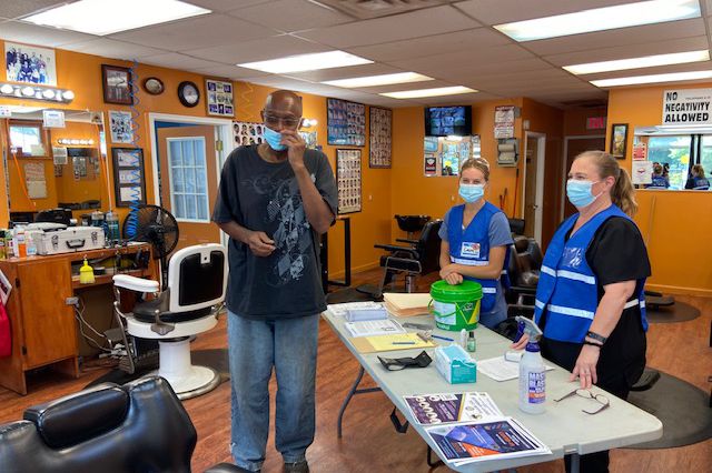 A man stands in front of a table with two healthcare workers. They are in a barbershop.